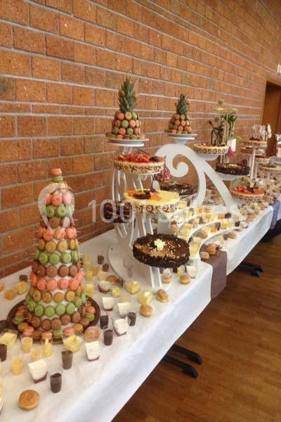 Table garnie de desserts variés, incluant des pyramides de macarons, gâteaux et verrines, dans une salle au mur en briques.