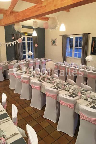 Salle décorée pour un événement avec des tables alignées, nappes blanches et accents roses, sous un plafond en bois.