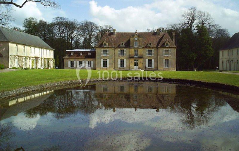 Manoir ancien en pierre avec toit en tuiles, entouré de pelouses et d'arbres, reflété dans un étang calme.