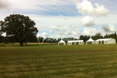 Miniature Location salle Brouay (Calvados) - Château de Brouay #14 Grand espace vert avec des tentes blanches installées sur une pelouse, entouré d'arbres et d'un ciel partiellement nuageux.