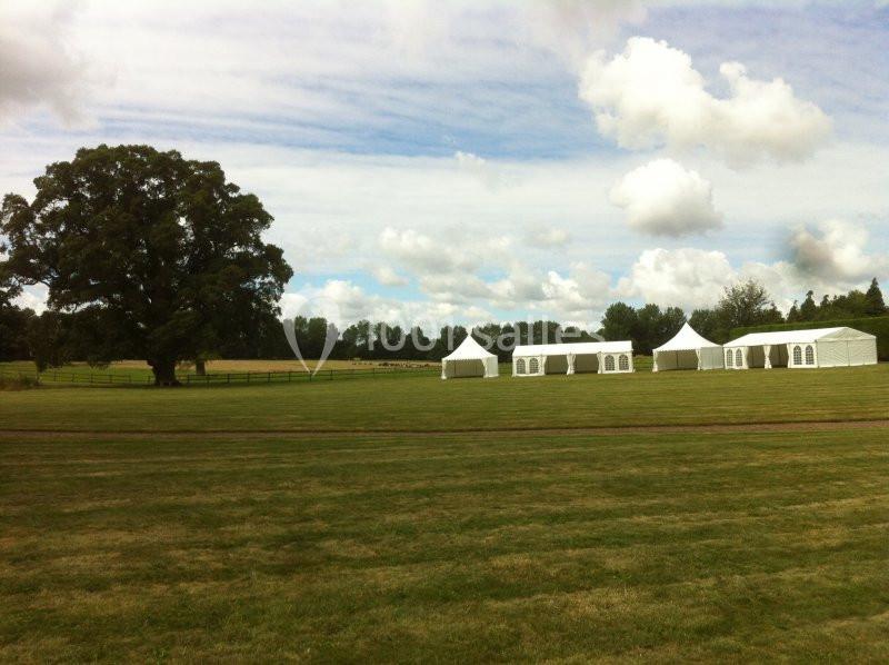 Location salle Brouay (Calvados) - Château de Brouay #14 Grand espace vert avec des tentes blanches installées sur une pelouse, entouré d'arbres et d'un ciel partiellement nuageux.