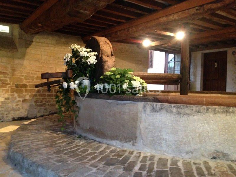 Location salle Brouay (Calvados) - Château de Brouay #13 Ancien pressoir en bois dans une salle en pierre, décoré de fleurs blanches et entouré de pavés.