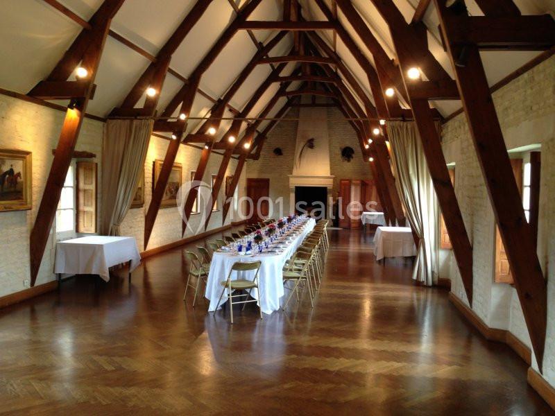 Location salle Brouay (Calvados) - Château de Brouay #4 Grande salle avec une longue table dressée pour un repas, entourée de chaises, sous une charpente en bois apparente.