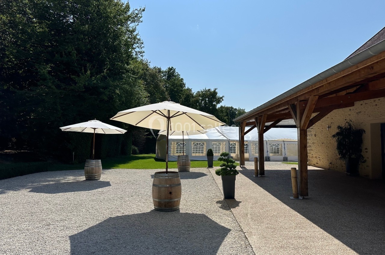 Cour extérieure avec parasols sur des tonneaux, espace gravillonné, pelouse et grandes tentes blanches en arrière-plan.