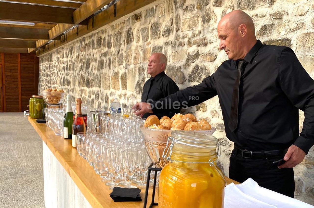 Deux serveurs en tenue noire préparent une table garnie de boissons, verrerie et condiments devant un mur en pierre.
