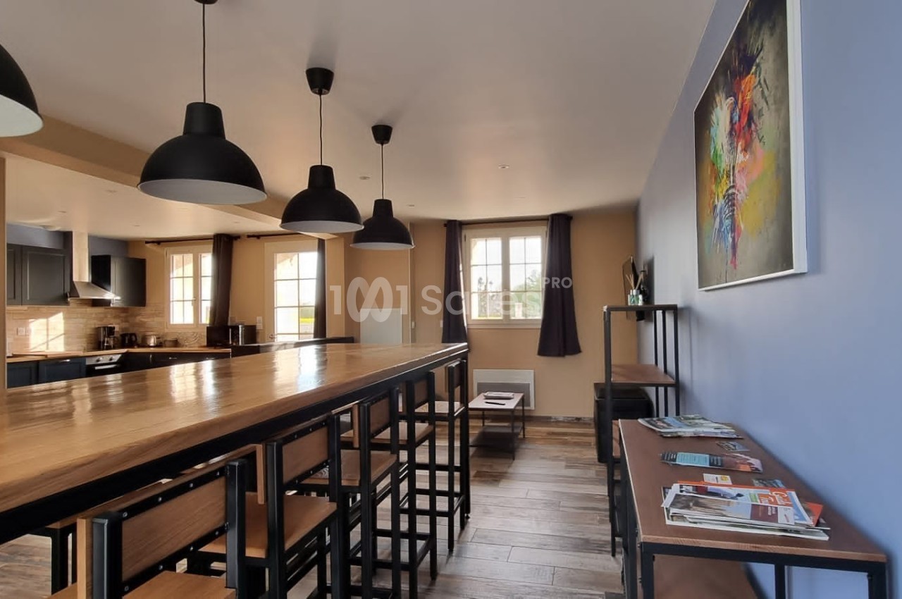 Salle à manger lumineuse avec une grande table en bois, des chaises hautes, des suspensions noires et un coin lecture.