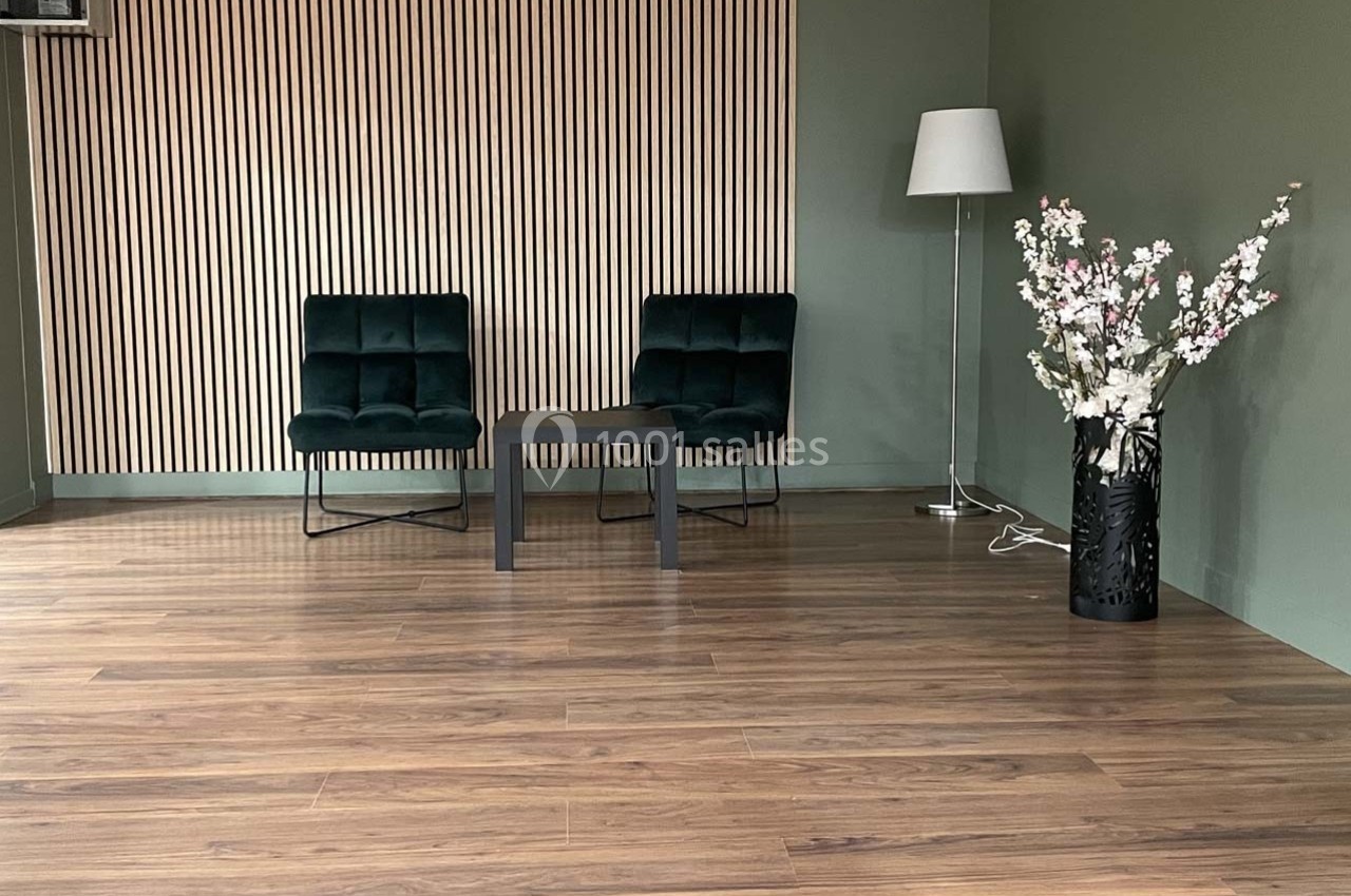 Deux fauteuils verts, une table basse, un lampadaire et un vase de fleurs dans une pièce au sol en bois et murs verts.