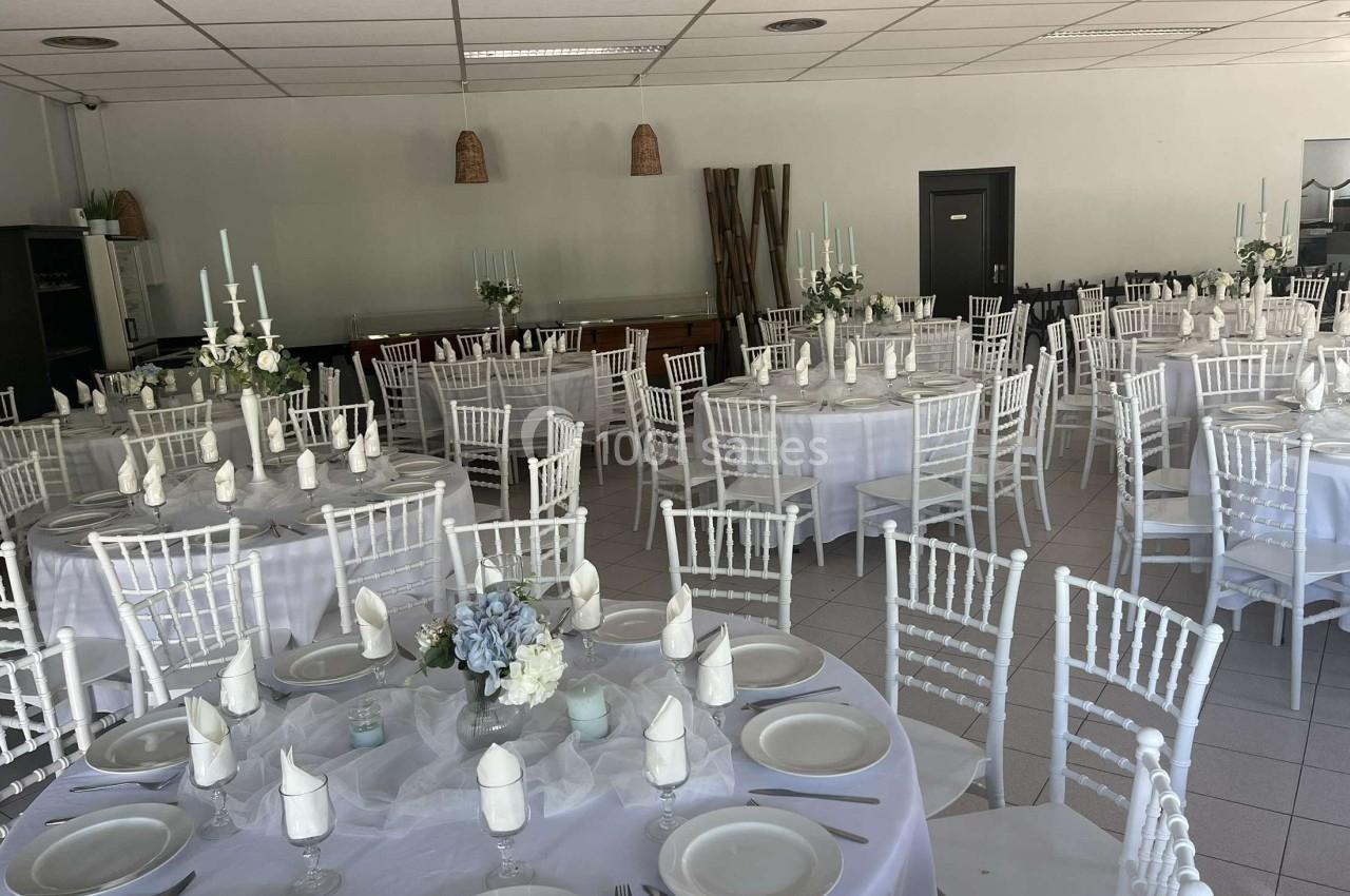 Salle de réception décorée avec des tables rondes dressées, nappes blanches, chaises blanches et centres de table floraux.