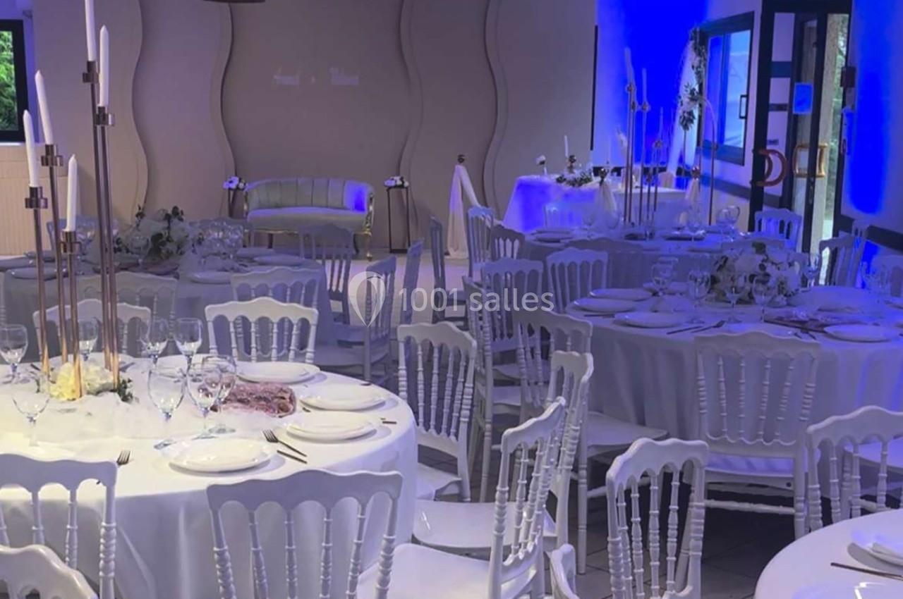 Salle de réception décorée avec des tables rondes dressées, chaises blanches et éclairage bleu en arrière-plan.