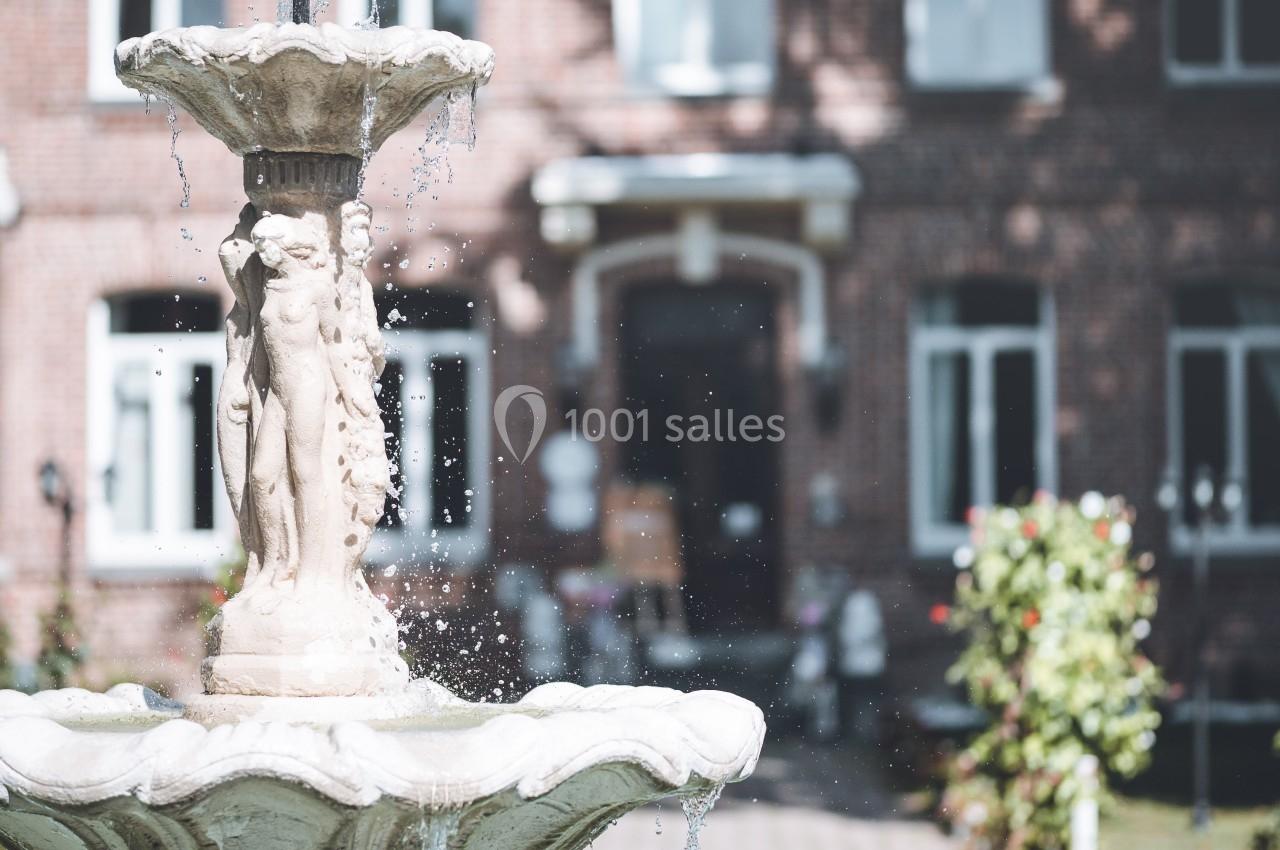 Fontaine en pierre ornée de sculptures, avec en arrière-plan une façade en briques et un jardin fleuri.