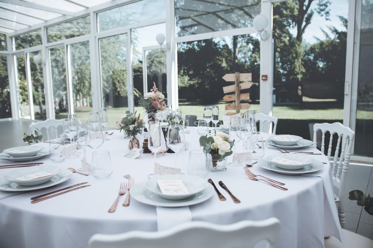 Table ronde élégamment dressée avec vaisselle blanche, décorations florales et vue sur un jardin à travers de grandes baies…