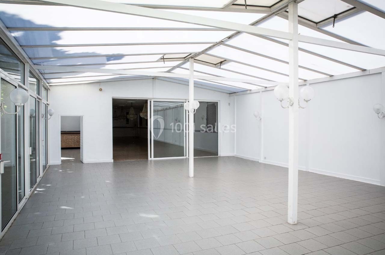 Salle lumineuse avec toit en verre, murs blancs et sol carrelé gris, donnant sur une pièce intérieure vitrée.