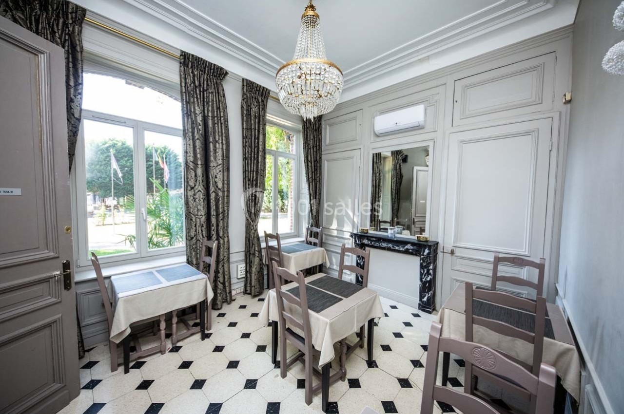 Salle lumineuse avec tables dressées, chaises en bois, sol en carrelage noir et blanc, grandes fenêtres et lustre élégant.
