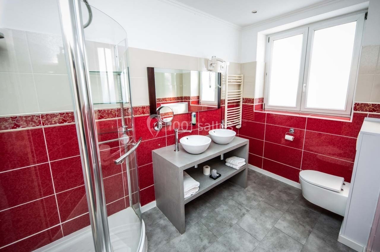 Salle de bain moderne avec douche vitrée, double vasque, carrelage rouge et blanc, et fenêtre apportant de la lumière…