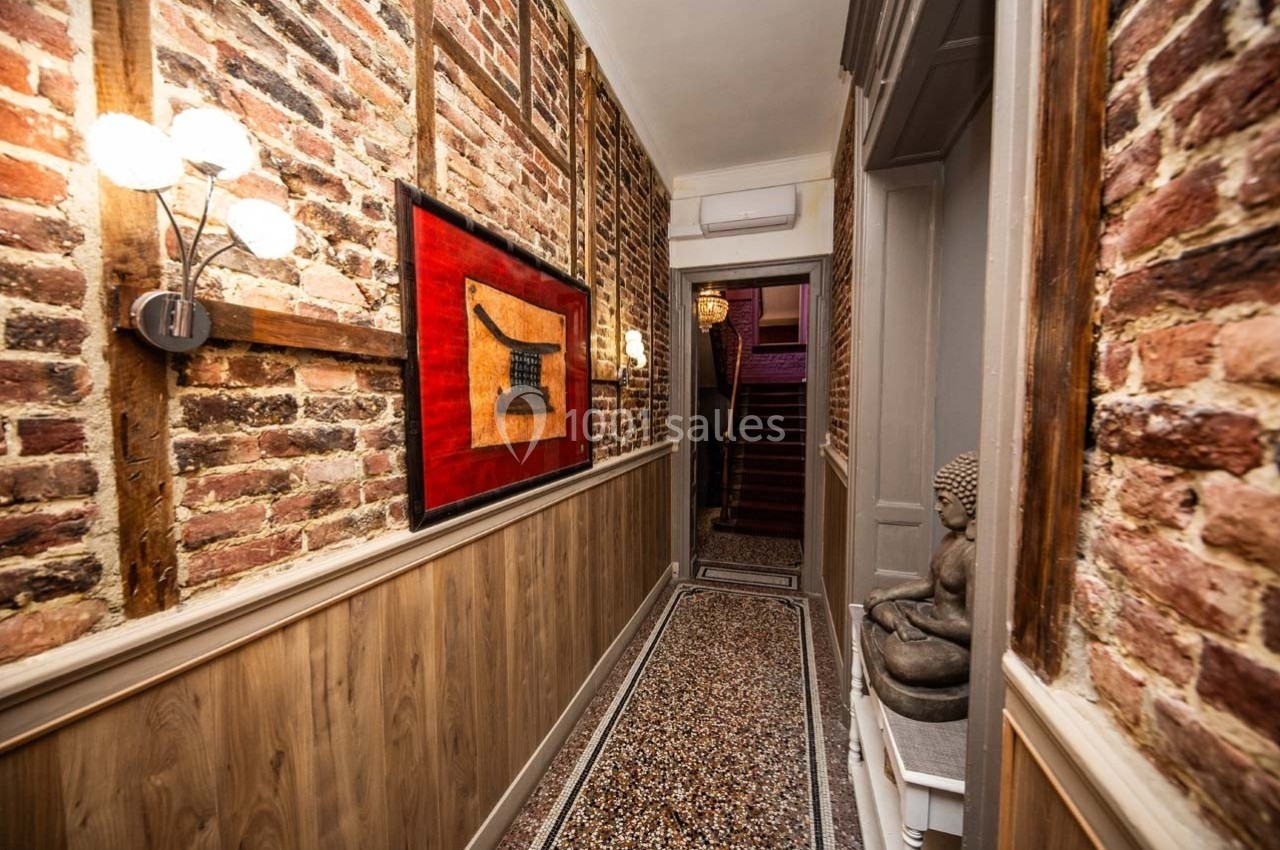 Couloir étroit avec murs en briques apparentes, tableau rouge, statue de Bouddha et escalier visible au fond.