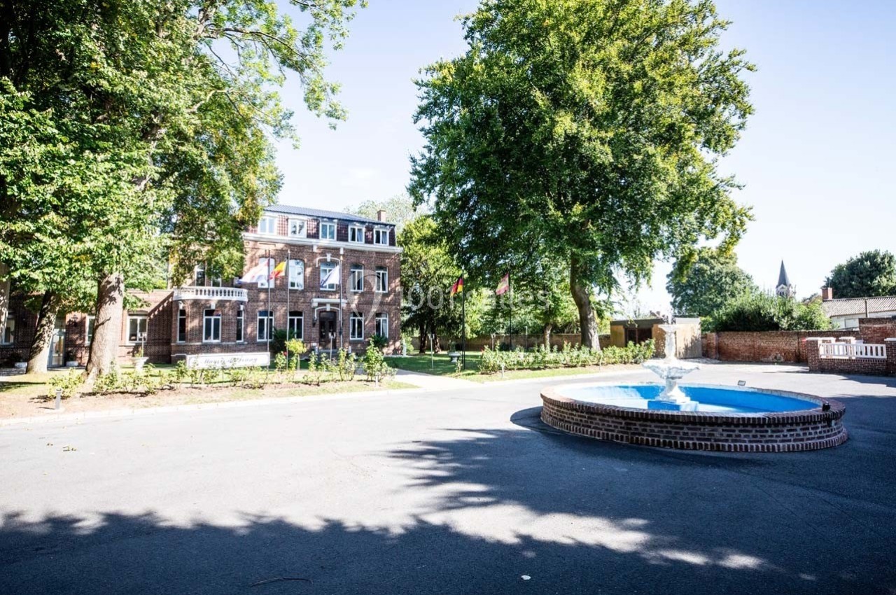 Bâtiment en briques entouré d'arbres avec une fontaine circulaire au premier plan, sous un ciel dégagé.