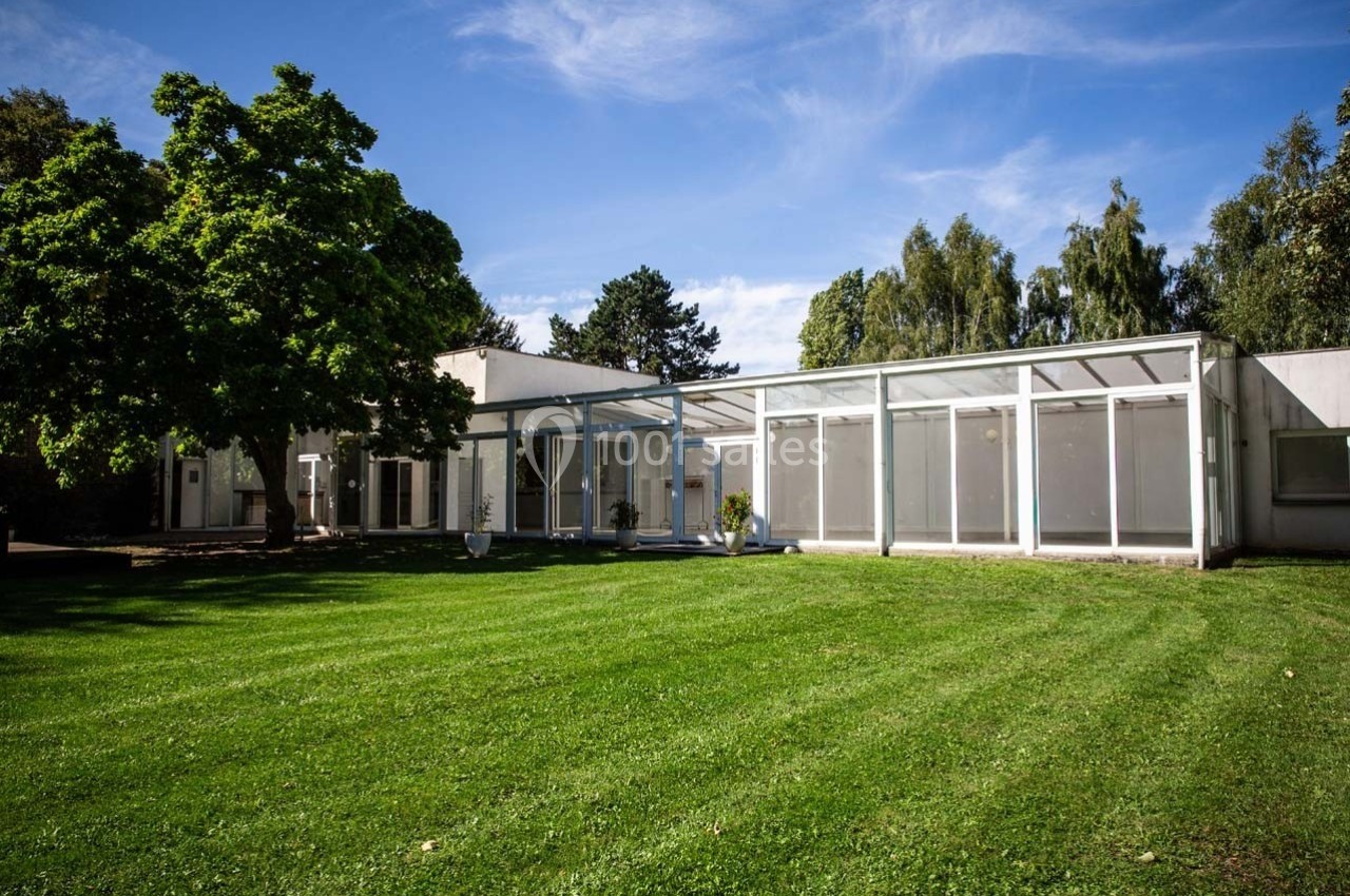 Bâtiment moderne avec une façade vitrée donnant sur une pelouse verte, entouré d'arbres sous un ciel bleu.