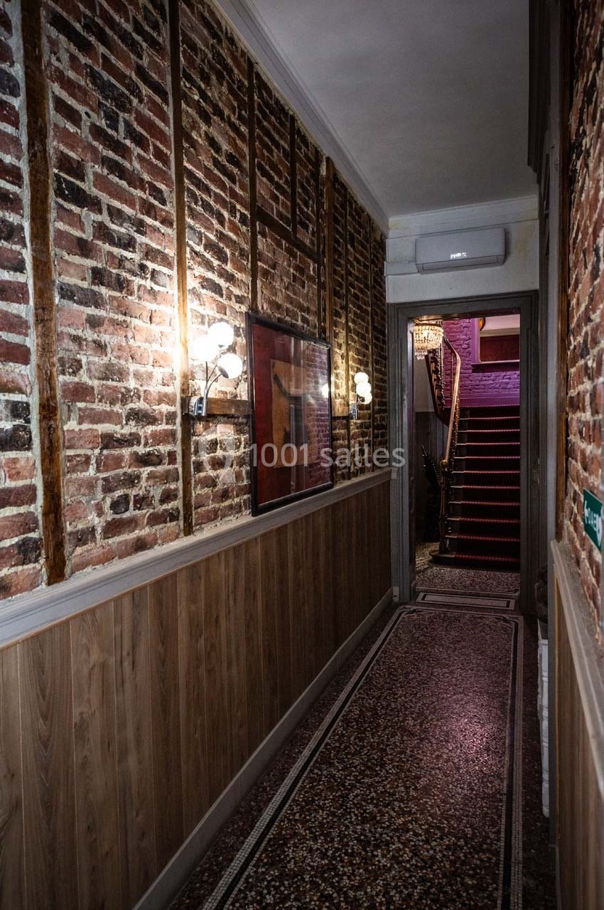 Couloir étroit avec murs en briques et bois, sol en terrazzo, éclairage mural et escalier au fond.
