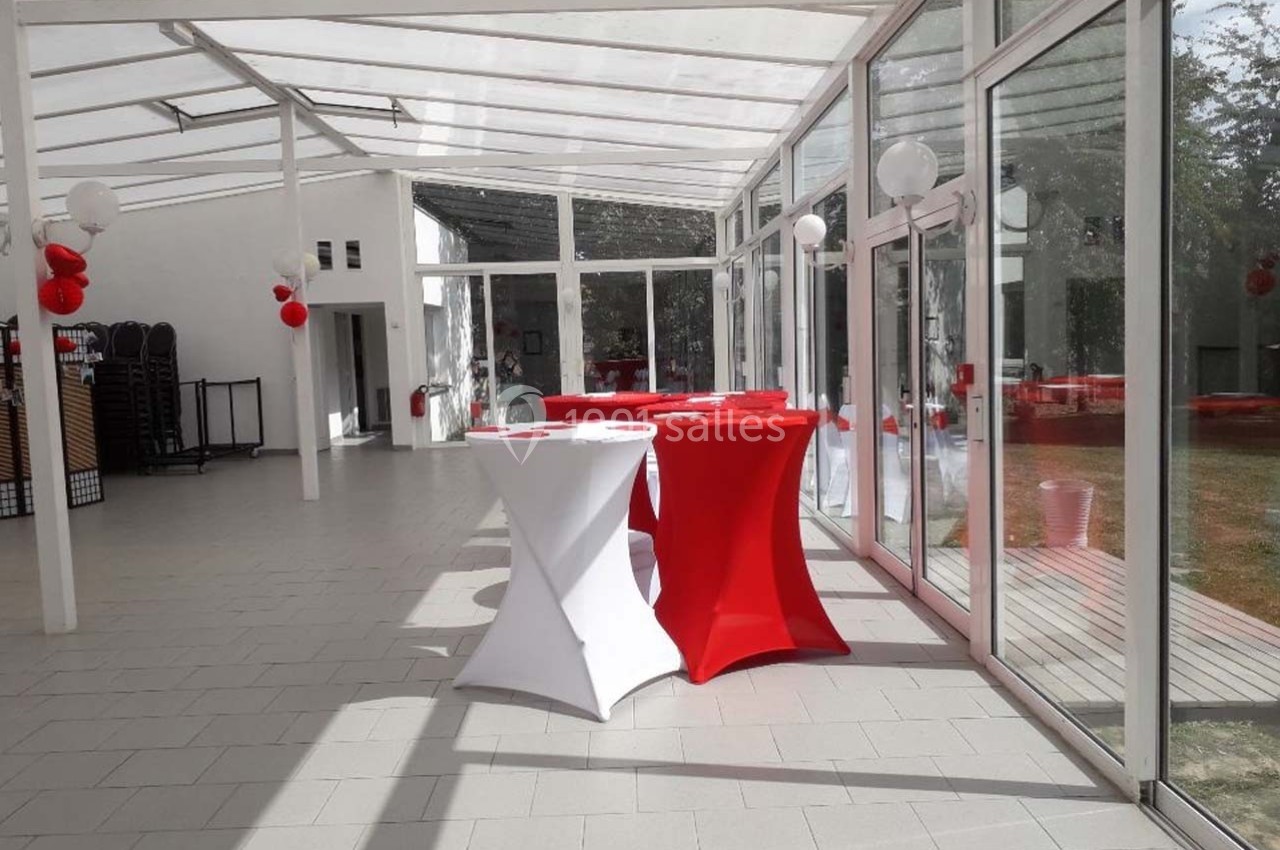 Salle lumineuse avec baies vitrées, tables hautes rouges et blanches décorées, ballons suspendus et vue sur l'extérieur.