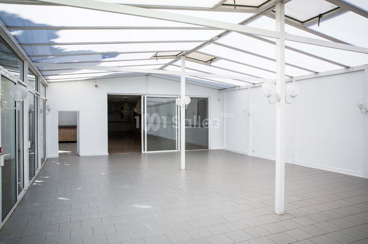 Salle lumineuse avec toit en verre, murs blancs et sol carrelé gris, donnant sur une pièce intérieure vitrée.