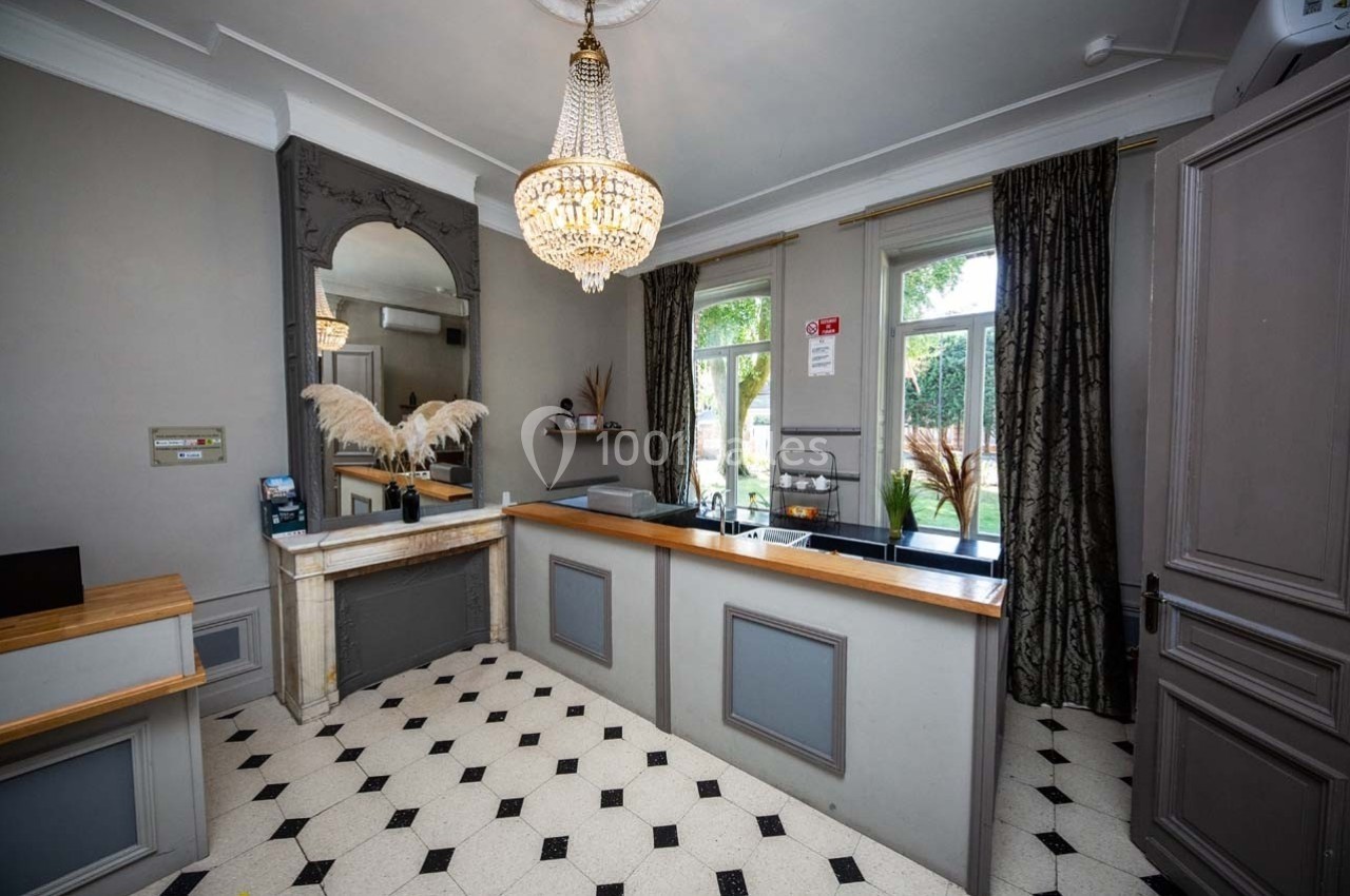 Réception d'un hôtel avec comptoir en bois, lustre élégant, miroir orné et sol carrelé noir et blanc.