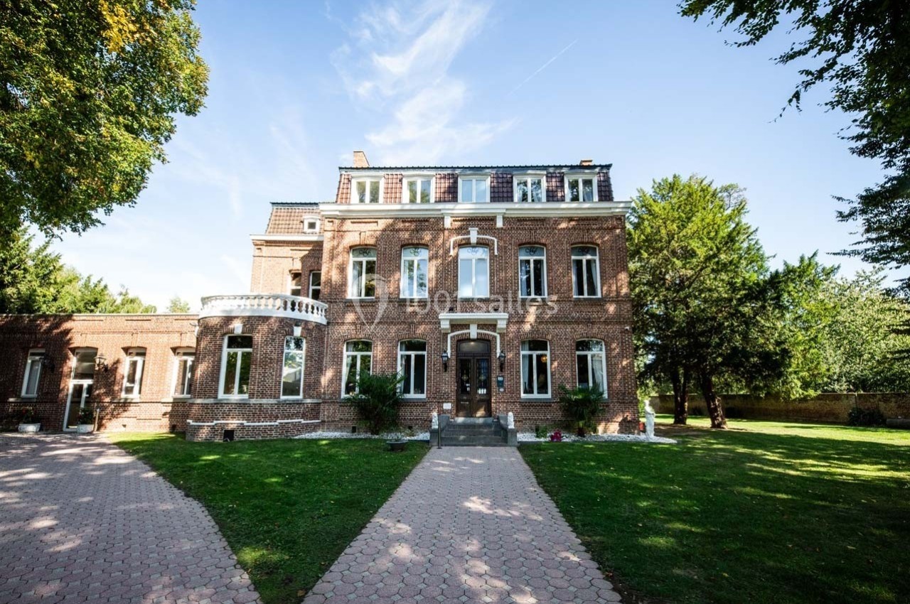 Grande maison en briques rouges avec toit mansardé, entourée d'un jardin arboré sous un ciel dégagé.