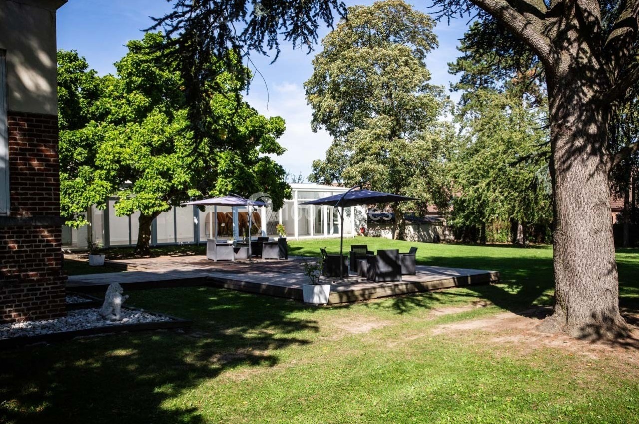 Terrasse aménagée avec tables et parasols dans un jardin arboré, entourée de pelouse et d'arbres.