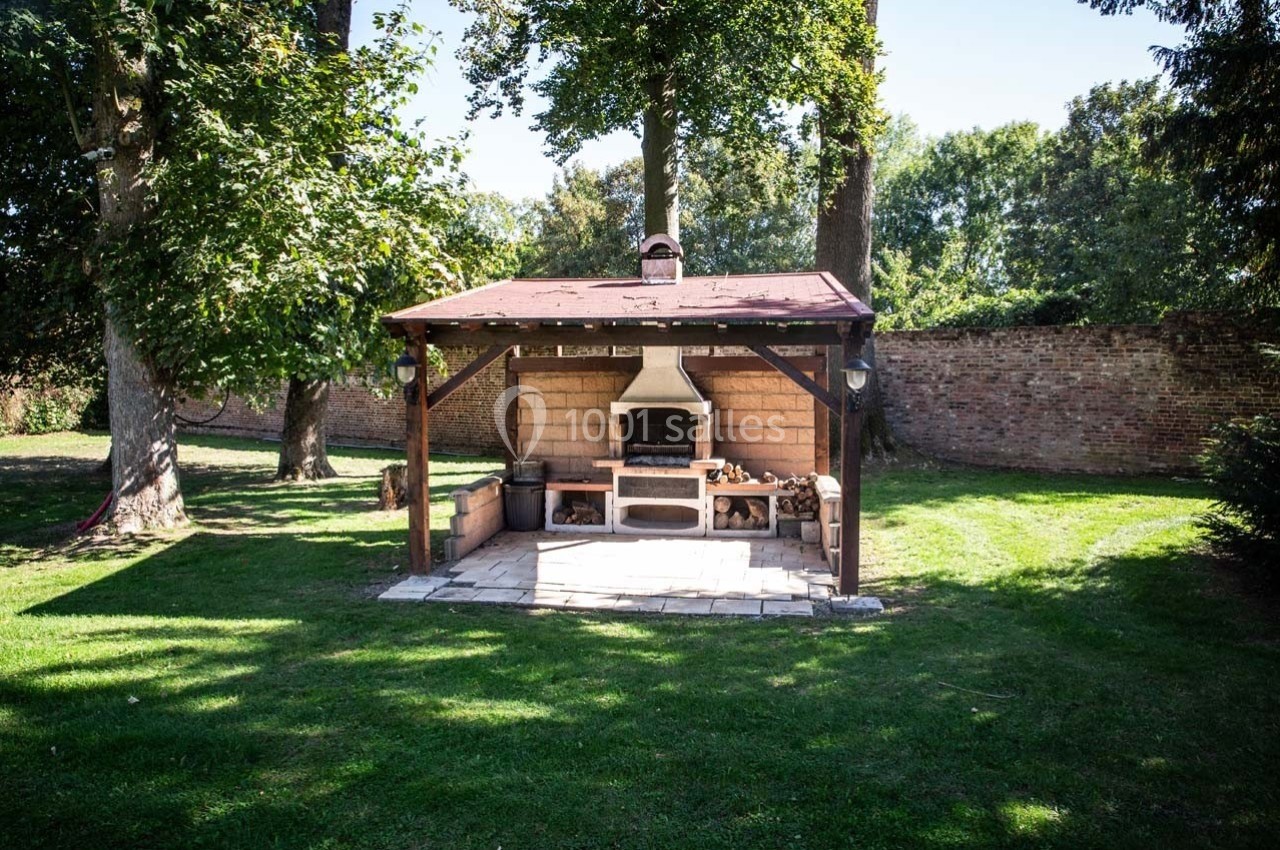 Abri de jardin en bois avec barbecue intégré, entouré de pelouse et d'arbres, devant un mur de briques.