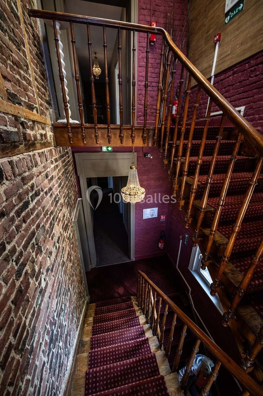 Escalier en bois avec tapis rouge, murs en briques apparentes et lustre suspendu dans un bâtiment ancien.