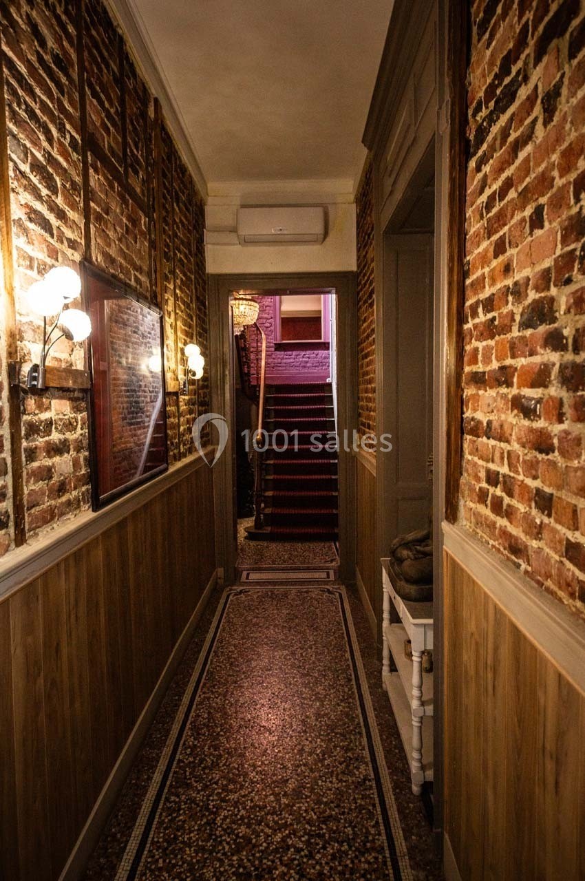 Couloir étroit avec murs en briques apparentes, sol en mosaïque, éclairage mural et escalier au fond.