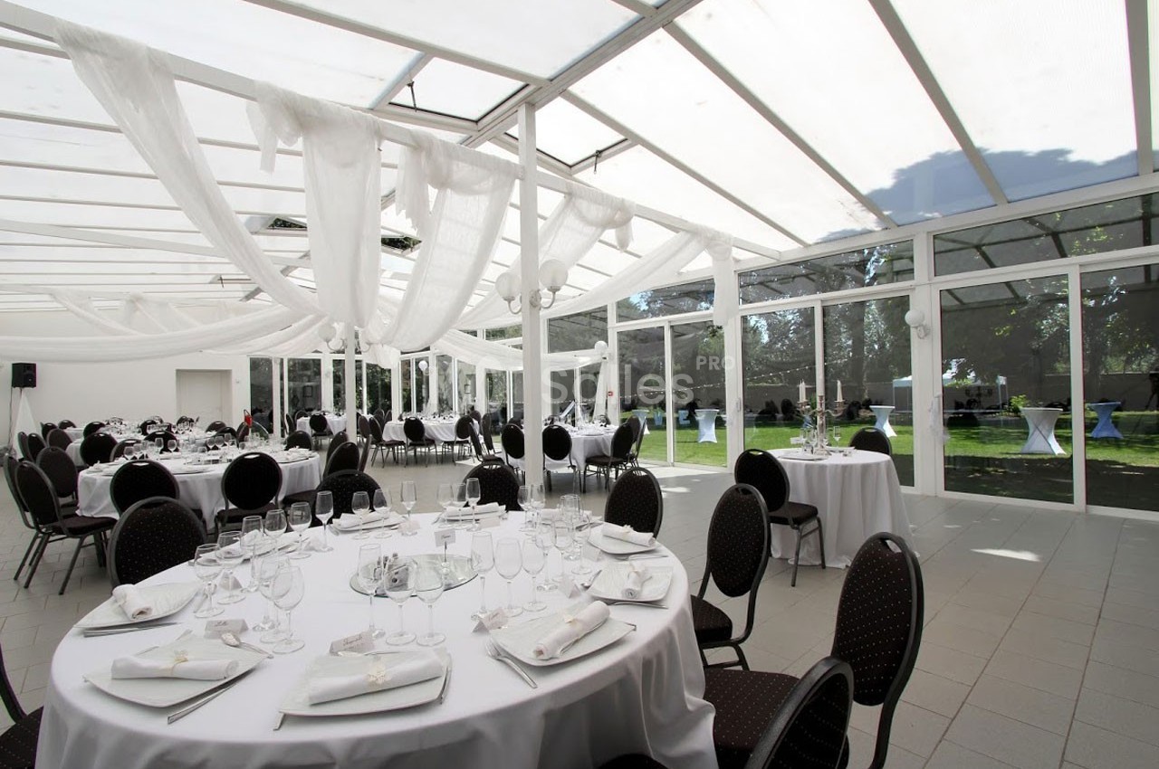 Salle de réception lumineuse avec tables rondes dressées, chaises noires et grandes baies vitrées donnant sur un jardin.