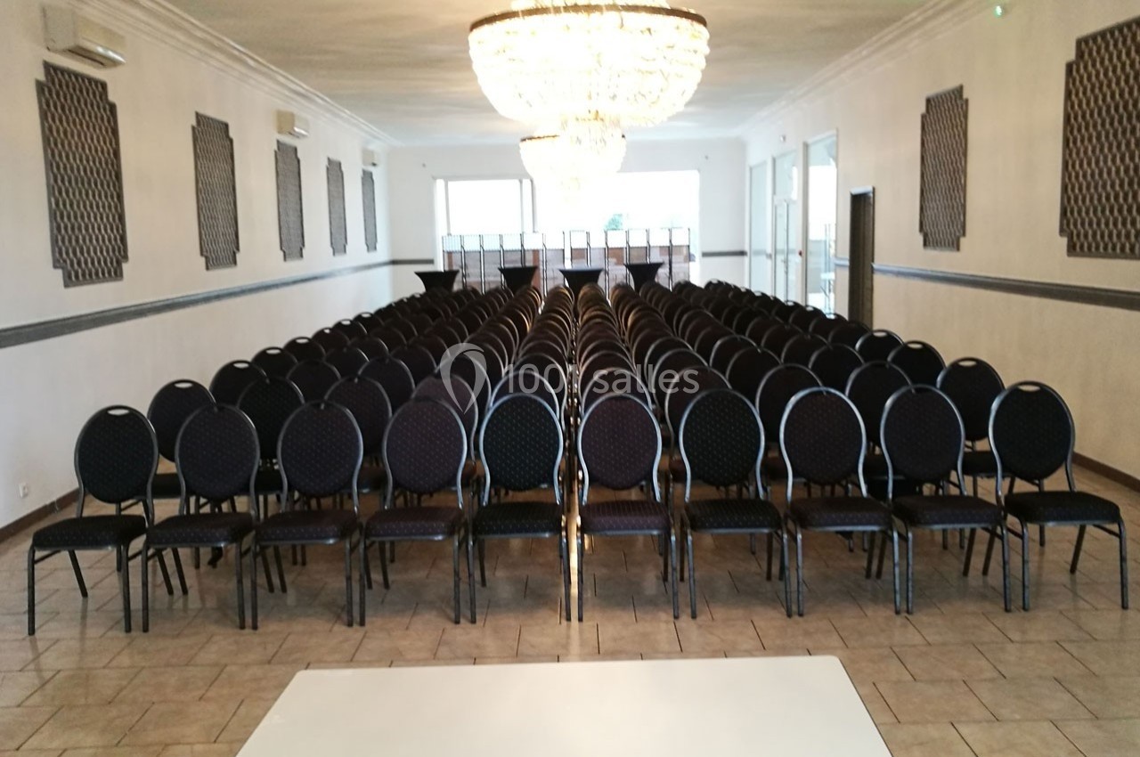 Salle de réception avec des rangées de chaises noires alignées face à une estrade, éclairée par un grand lustre.