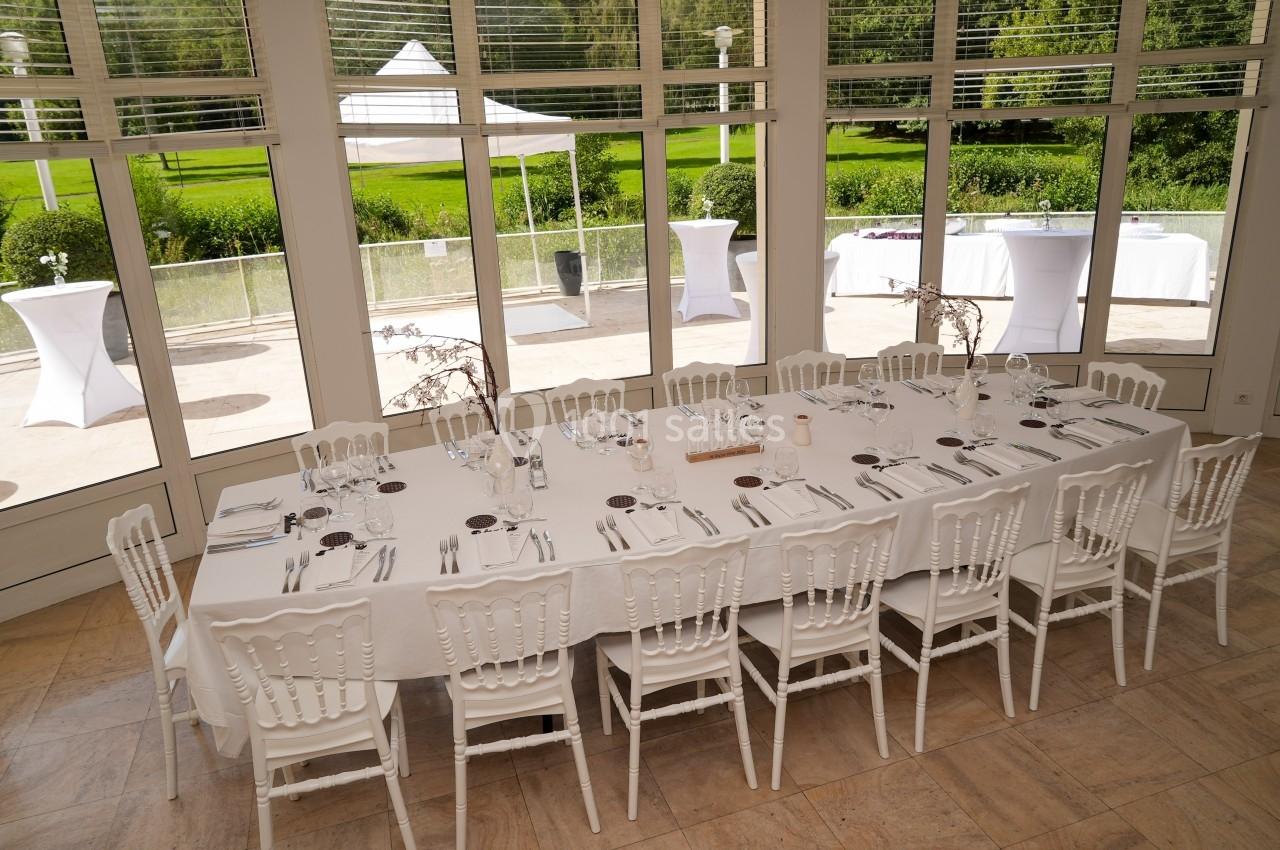Grande table rectangulaire dressée avec vaisselle blanche, entourée de chaises, dans une salle lumineuse avec vue sur un…