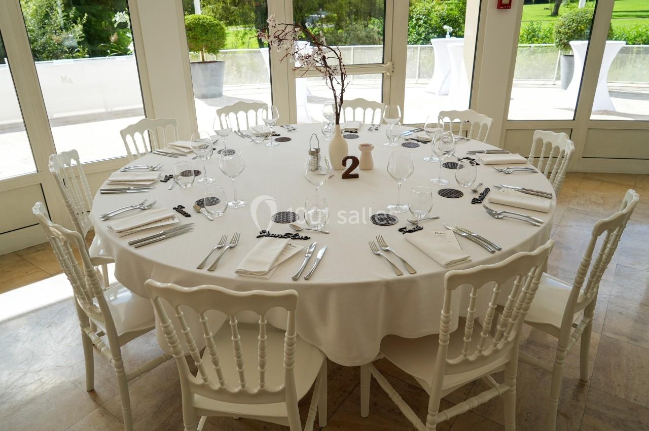 Table ronde dressée avec nappes blanches, couverts, verres et décorations, entourée de chaises blanches dans une salle…