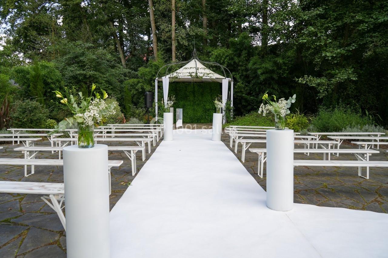 Allée centrale blanche menant à une arche de cérémonie en plein air, entourée de bancs et de végétation.