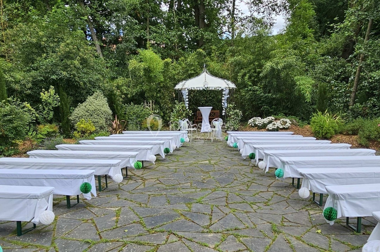 Allée centrale avec bancs recouverts de tissu blanc menant à une arche décorée, dans un jardin verdoyant.