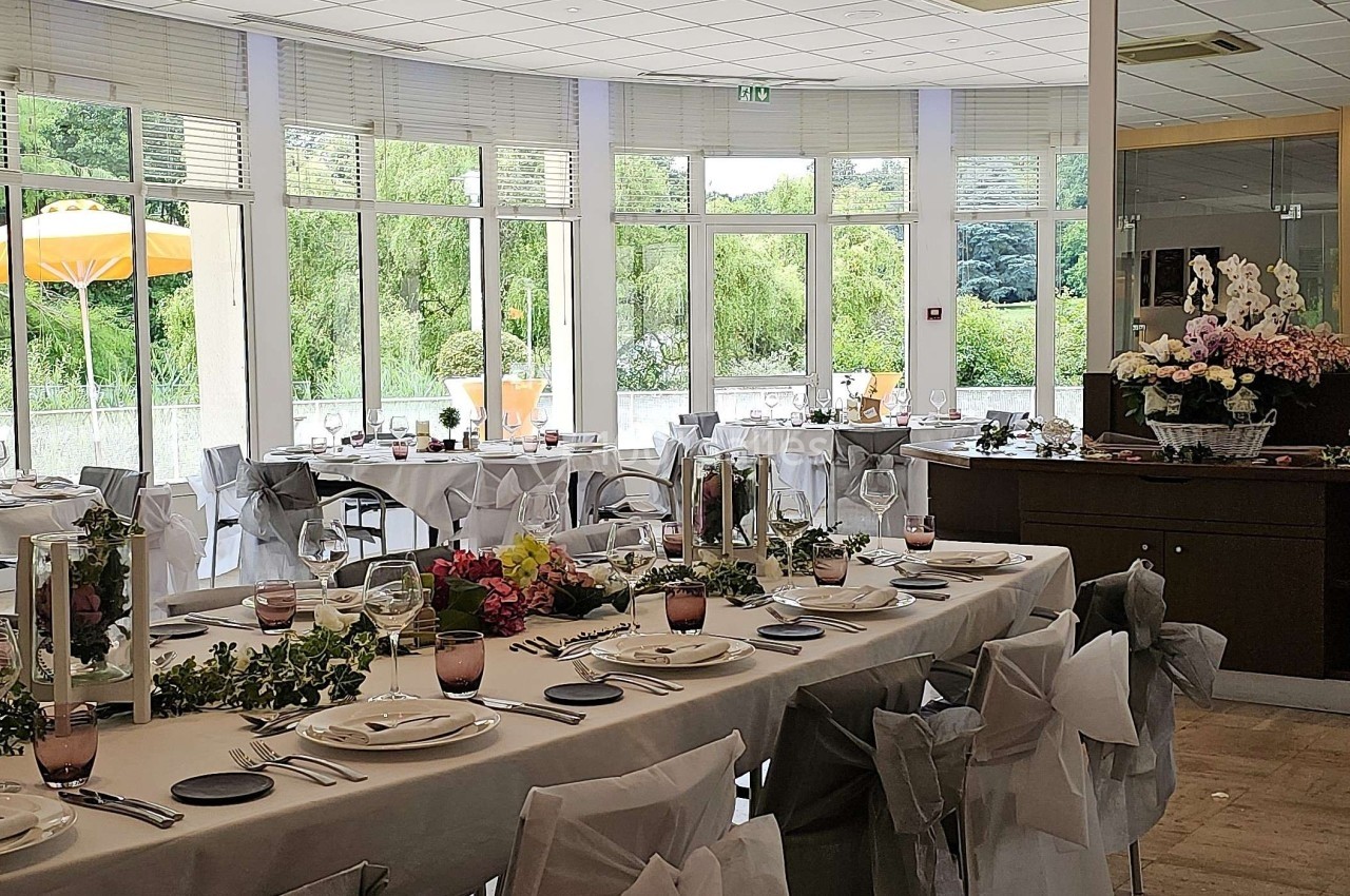 Salle de réception lumineuse avec tables décorées pour un événement, grandes baies vitrées donnant sur un jardin.