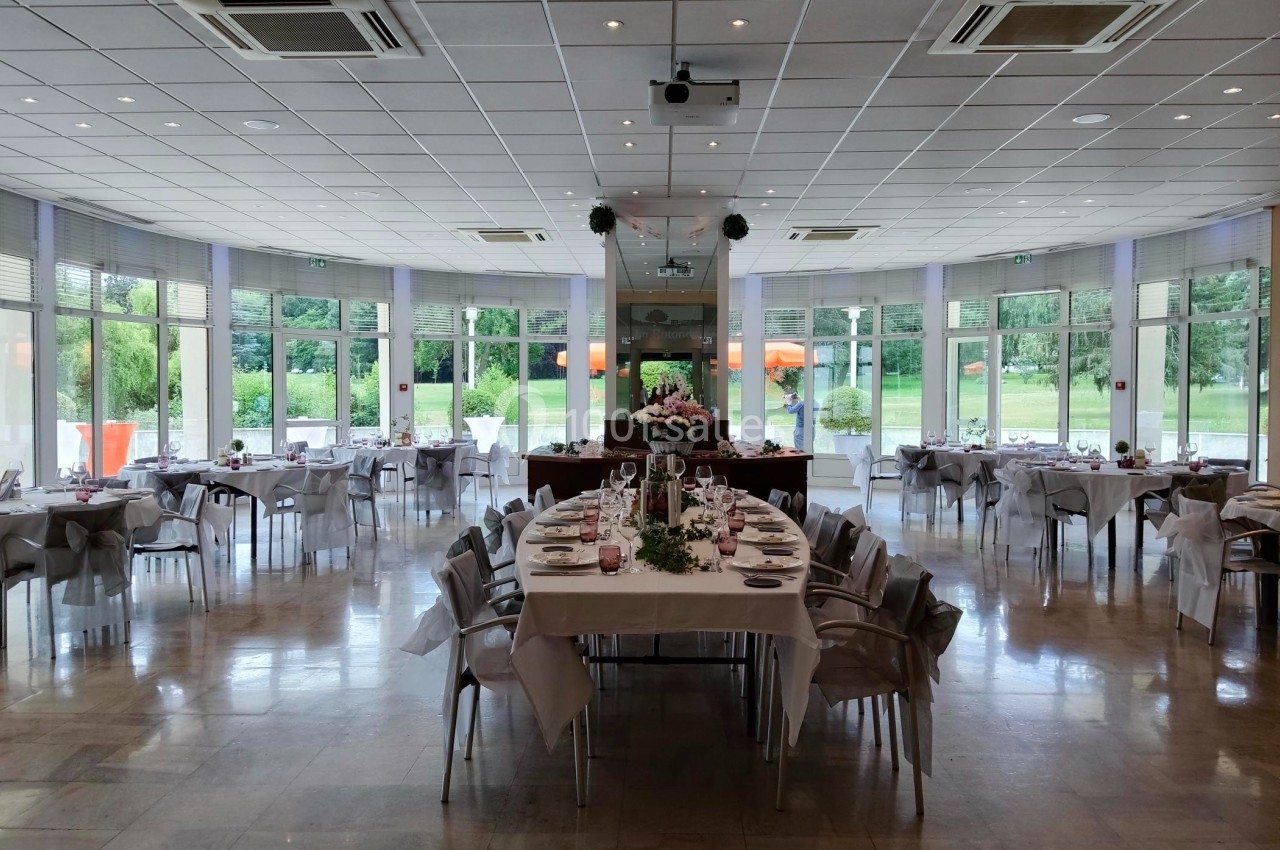 Salle de réception lumineuse avec tables dressées pour un événement, grandes baies vitrées donnant sur un jardin.