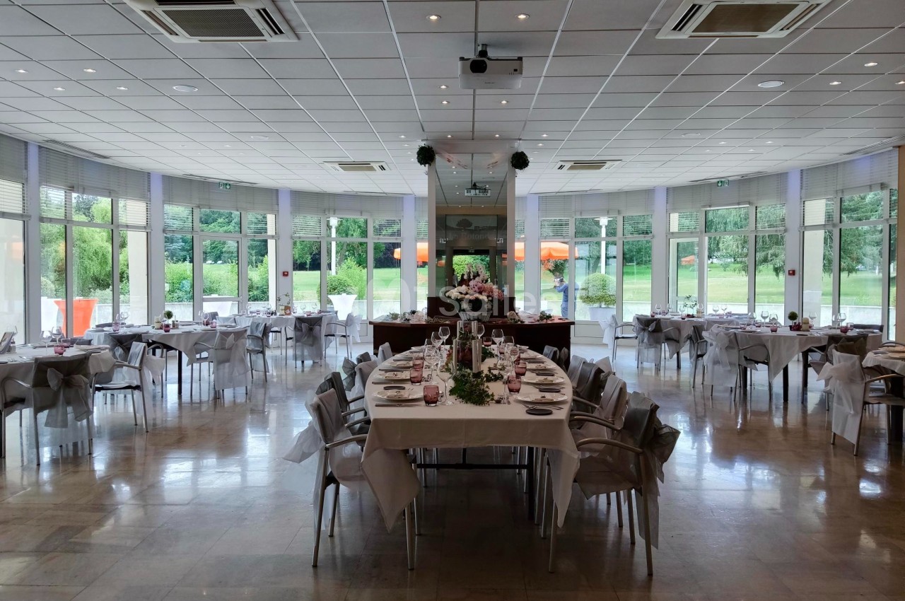 Salle de réception lumineuse avec tables dressées pour un événement, grandes baies vitrées donnant sur un jardin.