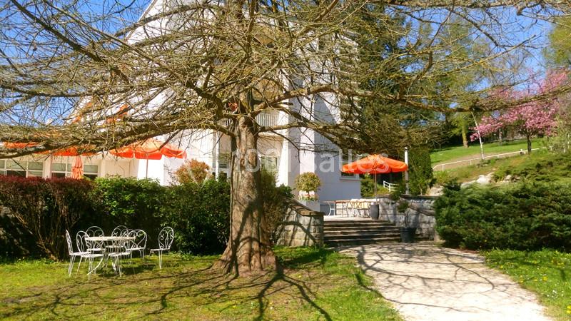 Maison blanche entourée d'arbres, avec terrasse équipée de parasols orange et mobilier de jardin en métal blanc.