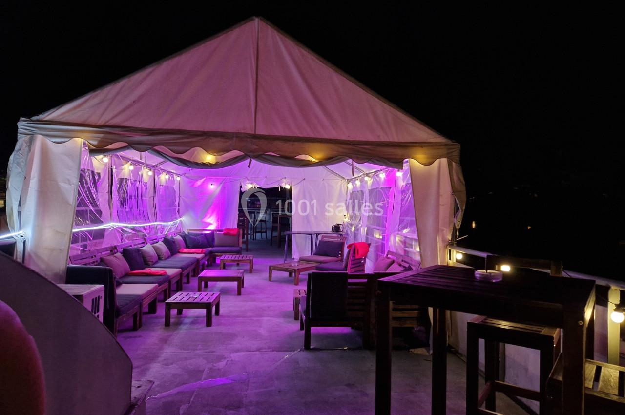 Tente éclairée de lumières violettes sur une terrasse, aménagée avec des fauteuils et des tables en soirée.