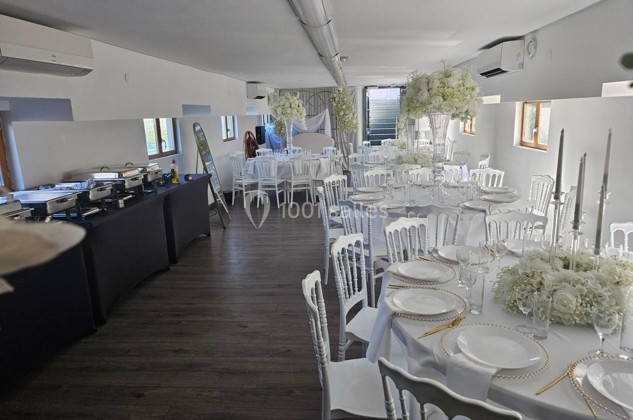 Salle de réception décorée avec des tables rondes dressées, chaises blanches et fleurs blanches suspendues.