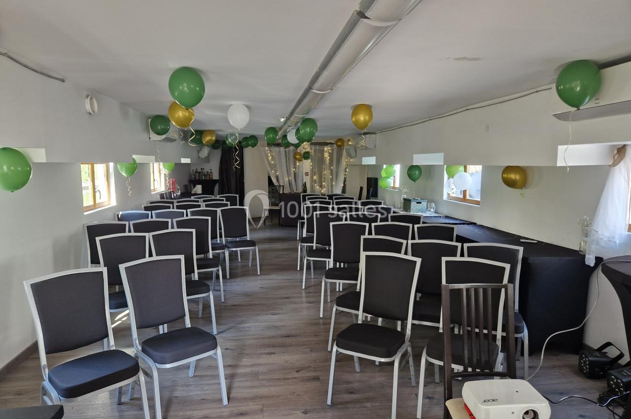 Salle aménagée avec des rangées de chaises noires, décorée de ballons verts, blancs et dorés, et équipée d'un projecteur.