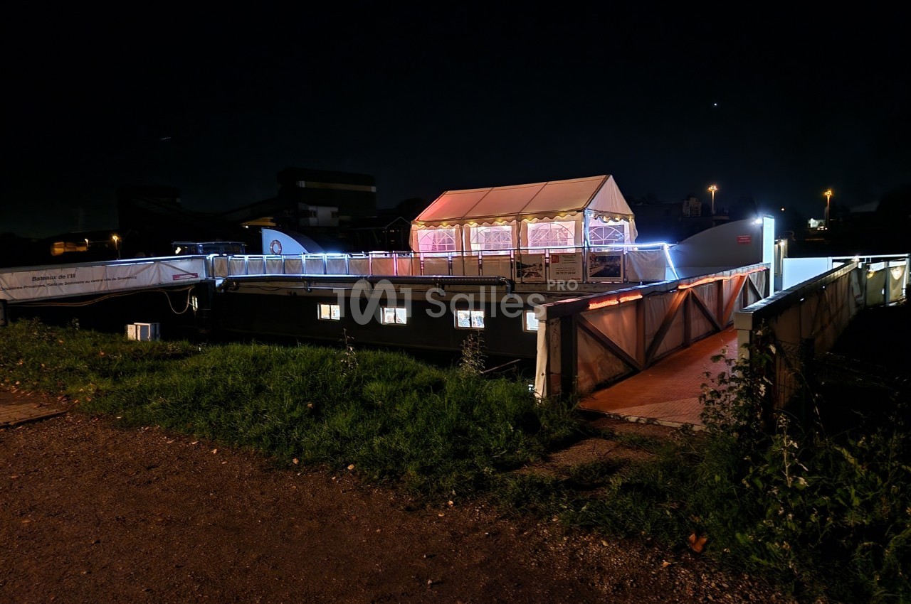 Bateau amarré de nuit avec une structure éclairée sur le pont, entouré d'herbe et de bâtiments en arrière-plan.