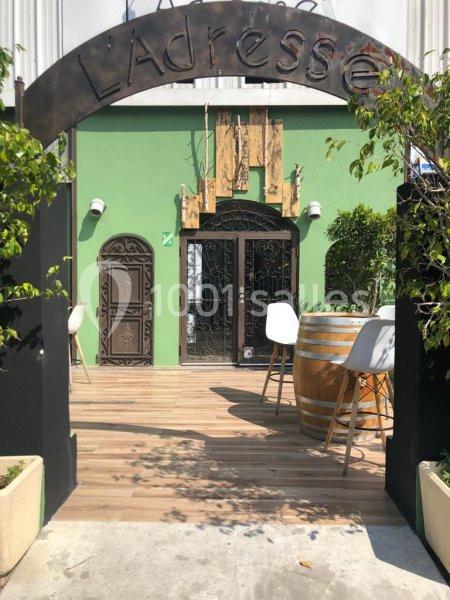 Entrée d'un café ou restaurant avec une arche en métal, terrasse en bois, tables hautes et façade verte.