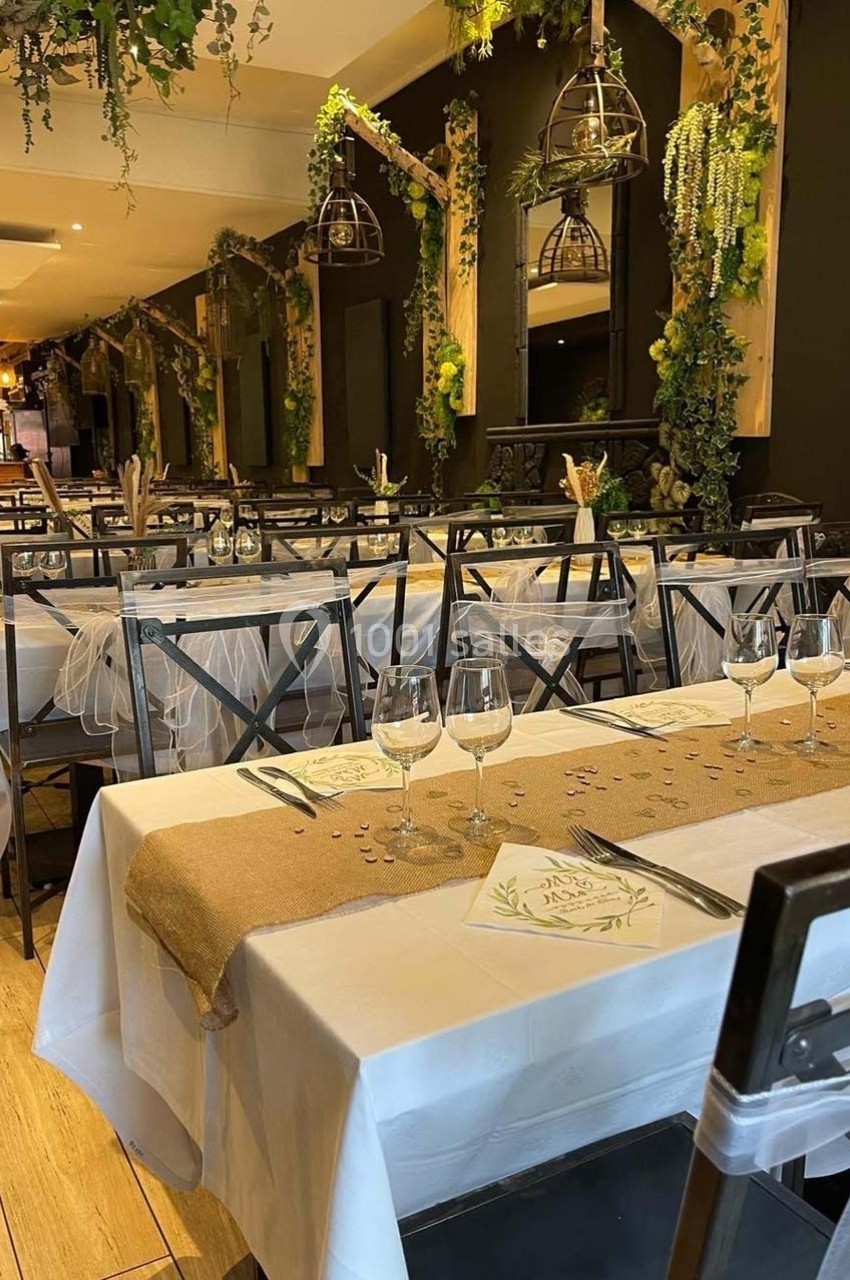 Salle de réception décorée avec des tables dressées, nappes blanches, chemins de table en toile de jute et plantes…
