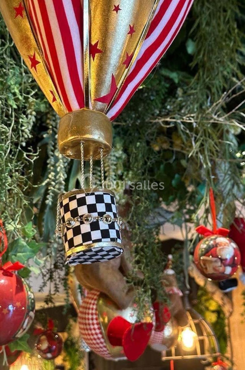 Montgolfière décorative suspendue avec motifs rayés et damiers, entourée de végétation et ornements festifs.
