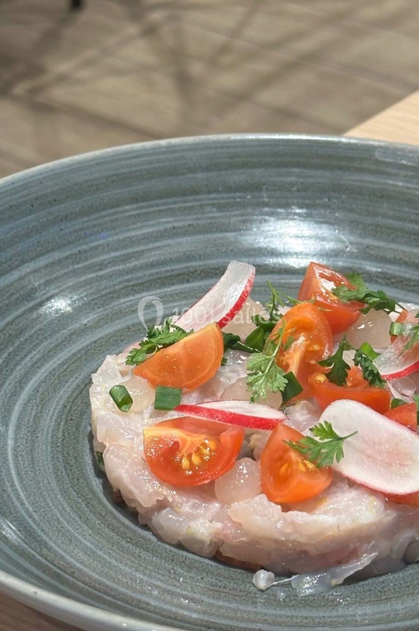 Tartare de poisson blanc garni de tomates cerises, radis et herbes fraîches, présenté dans une assiette grise.