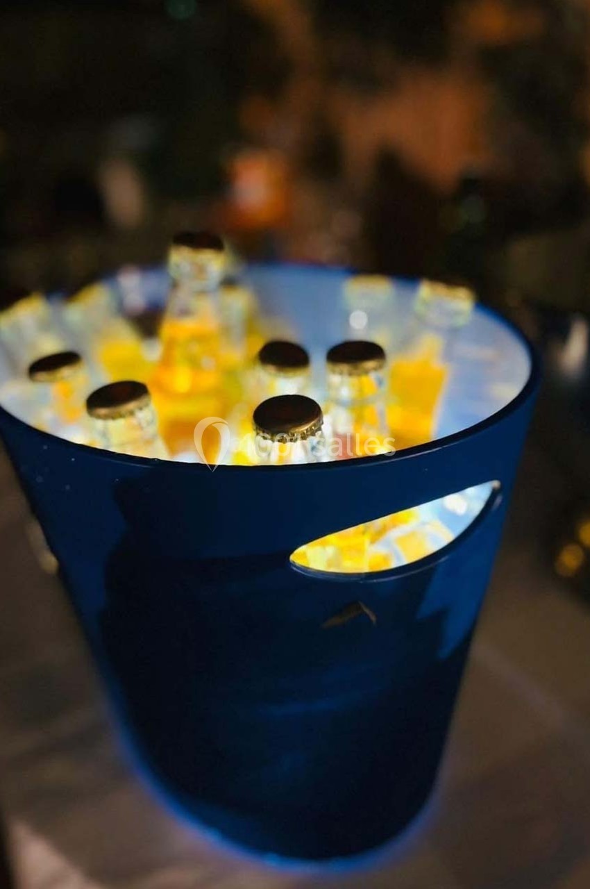 Seau bleu rempli de bouteilles en verre éclairées, posé sur une table dans un environnement faiblement éclairé.