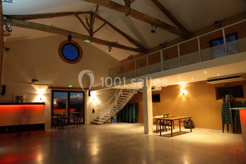 Salle spacieuse avec mezzanine, poutres apparentes, tables et chaises empilées, éclairage chaleureux.